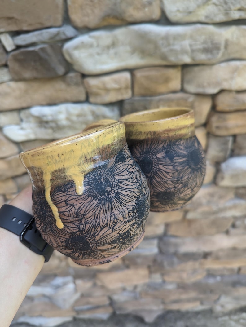 May include: Two ceramic mugs with a sunflower design. The mugs have a brown base with black sunflower patterns and a yellow rim. The glaze drips down the side of one mug. The background is a stone wall.