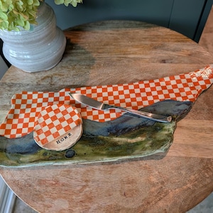 May include: A ceramic serving tray shaped like the state of Tennessee, featuring an orange and white checkered pattern on the top half and a mottled blue and green glaze on the bottom. A small round ceramic coaster with the word HOME is included.