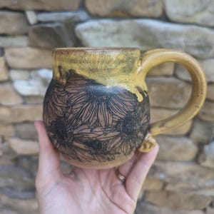 Puede incluir: Una taza de cerámica hecha a mano con un diseño de girasol. La taza tiene una base marrón con contornos de girasol negros y una parte superior y un asa amarillas. La taza está sostenida en la mano de una persona.