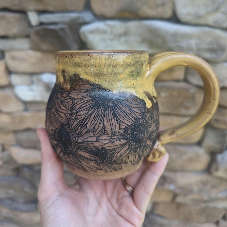 May include: A handmade ceramic mug with a sunflower design. The mug features a brown base with black sunflower outlines and a yellow top and handle. The mug is held in a person's hand.