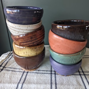 Puede incluir: Una pila de siete cuencos pequeños de cerámica hechos a mano en varios colores y patrones, incluyendo morado, blanco, marrón, amarillo y verde. Los cuencos están apilados sobre una superficie a cuadros blancos y negros.