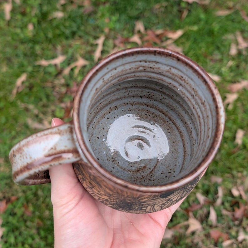 May include: Handmade ceramic mug with a brown exterior and a light blue interior. The mug features a detailed floral pattern around the base and a sturdy handle. The interior has a spiral design. The mug is perfect for coffee or tea.