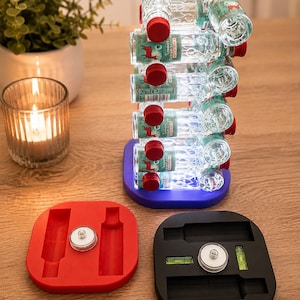 Könnte beinhalten: Ein vertikaler Ständer mit mehreren kleinen, durchsichtigen Glasflaschen mit roten Verschlüssen und Etiketten mit Hirschmotiv, gestapelt auf einem blauen Sockel. Zwei weitere quadratische Sockel in Rot und Schwarz sind ebenfalls abgebildet.