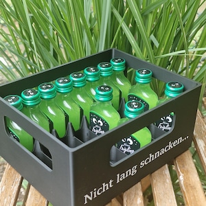 Könnte beinhalten: Eine schwarze Kiste gefüllt mit kleinen grünen Flaschen mit grünen Deckeln. Die Flaschen enthalten eine grüne Flüssigkeit und tragen ein Etikett mit der Aufschrift "Feigling". Auf der Seite der Kiste steht "Nicht lang schnacken...".