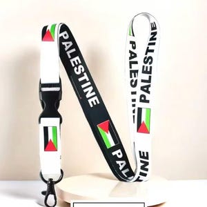 Puede incluir: Un cordón con la palabra "PALESTINA" impresa en blanco sobre fondo negro y blanco. El cordón presenta la bandera palestina y un cierre negro con un gancho metálico.