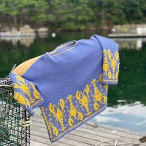 May include: A blue knitted sweater featuring yellow lobster motifs. The garment is displayed over a lobster pot on a wooden pier. The background shows water and trees.