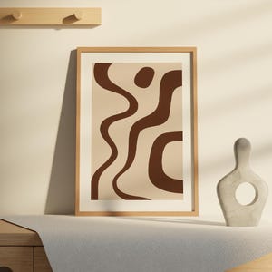May include: A framed abstract art print featuring brown organic shapes on a beige background. The artwork is displayed on a table next to a decorative sculpture. The frame is made of light-colored wood.
