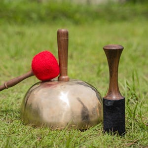 Op de afbeelding: Een bronzen zangkom met een houten hamer en een rode vilten klopper. Er is ook een tweede houten hamer met een zwarte vilten basis. De items staan op groen gras.