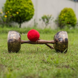 May include: A set of bronze-coloured singing bowls with a wooden handle and a red felt mallet, set on green grass. The bowls have a shiny, textured surface. The background features green foliage and a white wall.