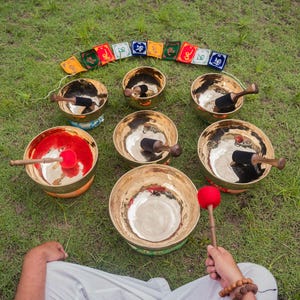 May include: A collection of brass singing bowls of varying sizes, arranged on green grass. Each bowl has a wooden mallet, and some have a red or black interior. Colorful prayer flags are arranged in an arc above the bowls.