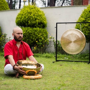 May include: A man in a red shirt and white pants sits on the grass, holding a golden singing bowl. A large gong hangs on a black stand nearby. The scene is set outdoors with green grass and bushes.