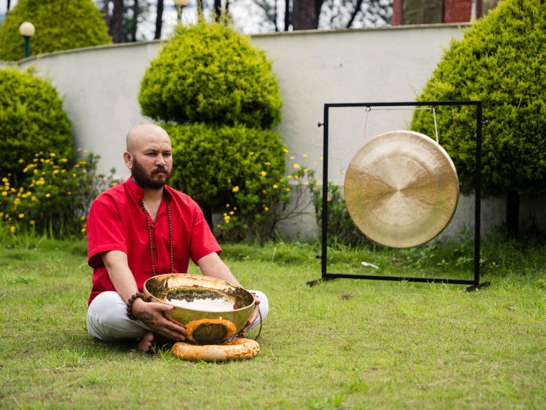 May include: A man in a red shirt and white pants sits on the grass, holding a golden singing bowl. A large gong hangs on a black stand nearby. The scene is set outdoors with green grass and bushes.