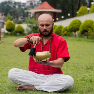 May include: A man in a red shirt and white pants sits cross-legged on green grass, holding a golden singing bowl. He is using a black mallet to strike the bowl. The background includes trees and a small red-roofed structure.