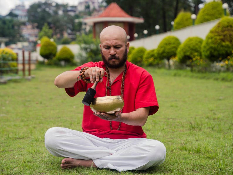 May include: A man in a red shirt and white pants sits cross-legged on green grass, holding a golden singing bowl. He is using a black mallet to strike the bowl. The background includes trees and a small red-roofed structure.