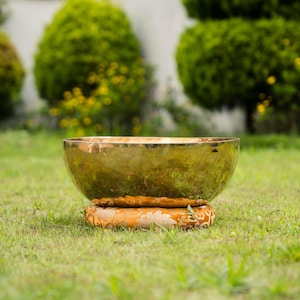 May include: A golden singing bowl with a wide, rounded shape, resting on a patterned orange cushion. The bowl is set on a bed of green grass, with blurred green foliage in the background.