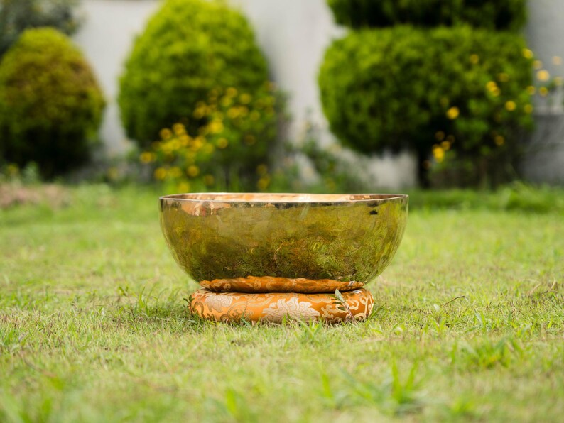 May include: A golden singing bowl with a wide, rounded shape, resting on a patterned orange cushion. The bowl is set on a bed of green grass, with blurred green foliage in the background.