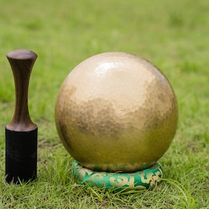 May include: A golden singing bowl with a textured surface, resting on a green cushion with gold accents. A wooden mallet with a black base stands to the left. The objects are set on a bed of green grass.