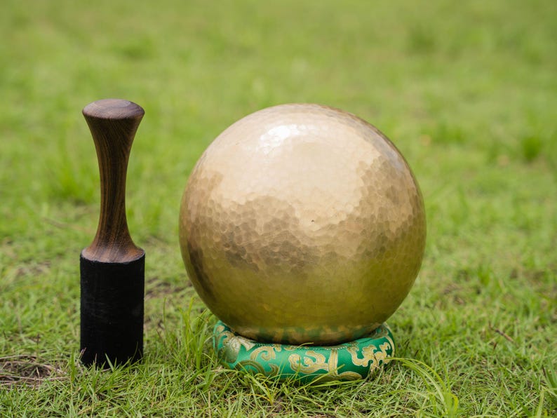 May include: A golden singing bowl with a textured surface, resting on a green cushion with gold accents. A wooden mallet with a black base stands to the left. The objects are set on a bed of green grass.