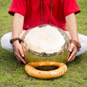 May include: A large, golden-colored gong with a circular, orange-colored cushion. The gong is held by a person wearing a red shirt and white pants, sitting on green grass. The gong is a musical instrument.