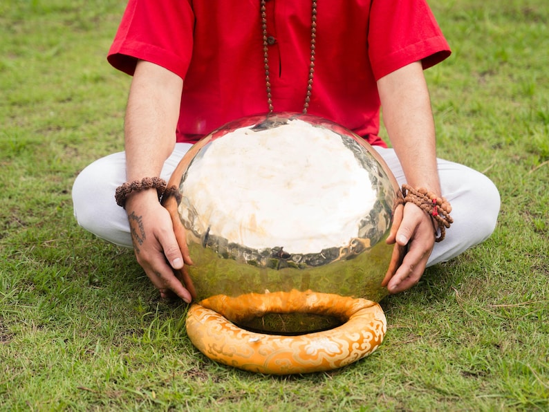 May include: A large, golden-colored gong with a circular, orange-colored cushion. The gong is held by a person wearing a red shirt and white pants, sitting on green grass. The gong is a musical instrument.