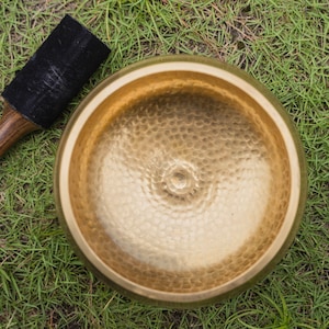 May include: A golden singing bowl with a textured surface and a wooden mallet with a black suede striking end. The bowl and mallet are resting on a bed of green grass. The bowl is a meditation tool.