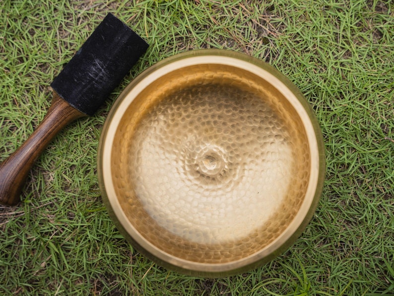 May include: A golden singing bowl with a textured surface and a wooden mallet with a black suede striking end. The bowl and mallet are resting on a bed of green grass. The bowl is a meditation tool.
