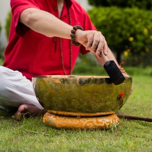 May include: A person seated on the grass is using a mallet to play a large, antique-looking singing bowl. The bowl is gold-colored and rests on a decorative, patterned cushion. A red mallet and a red ball are also visible.