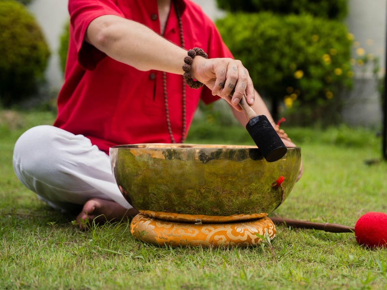May include: A person seated on the grass is using a mallet to play a large, antique-looking singing bowl. The bowl is gold-colored and rests on a decorative, patterned cushion. A red mallet and a red ball are also visible.