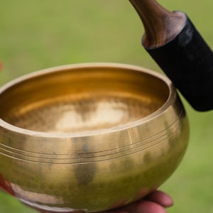 May include: A golden singing bowl with three etched lines near the rim. A wooden mallet with a black felt striking end rests nearby. The bowl is held against a blurred green background.