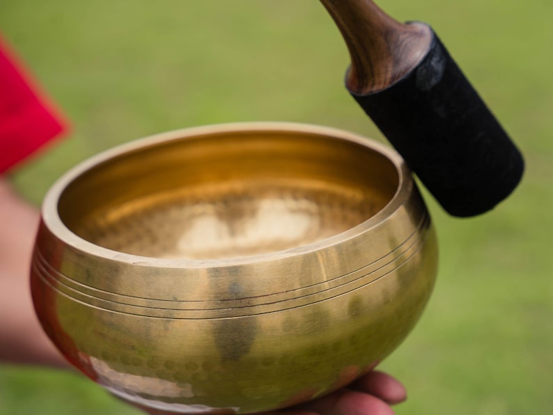 May include: A golden singing bowl with three etched lines near the rim. A wooden mallet with a black felt striking end rests nearby. The bowl is held against a blurred green background.