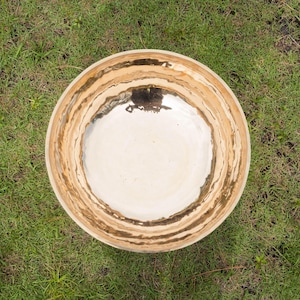 May include: Overhead view of a large, round ceramic bowl with a cream-colored interior and a textured, brown and tan rim. The bowl sits on a bed of green grass, creating a natural, earthy aesthetic.