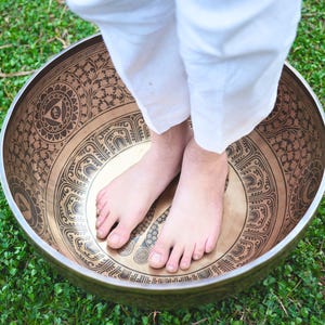 May include: A large, bronze-coloured singing bowl with intricate etched patterns. The bowl is round and has a wide rim. Bare feet are inside the bowl, and the background is green grass.