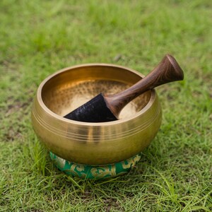 May include: A golden singing bowl with a wooden mallet, resting on a green and gold patterned cushion. The bowl is set against a backdrop of green grass, suggesting a focus on meditation and sound healing.