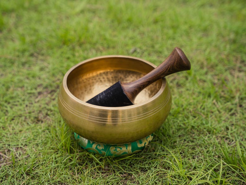 May include: A golden singing bowl with a wooden mallet, resting on a green and gold patterned cushion. The bowl is set against a backdrop of green grass, suggesting a focus on meditation and sound healing.