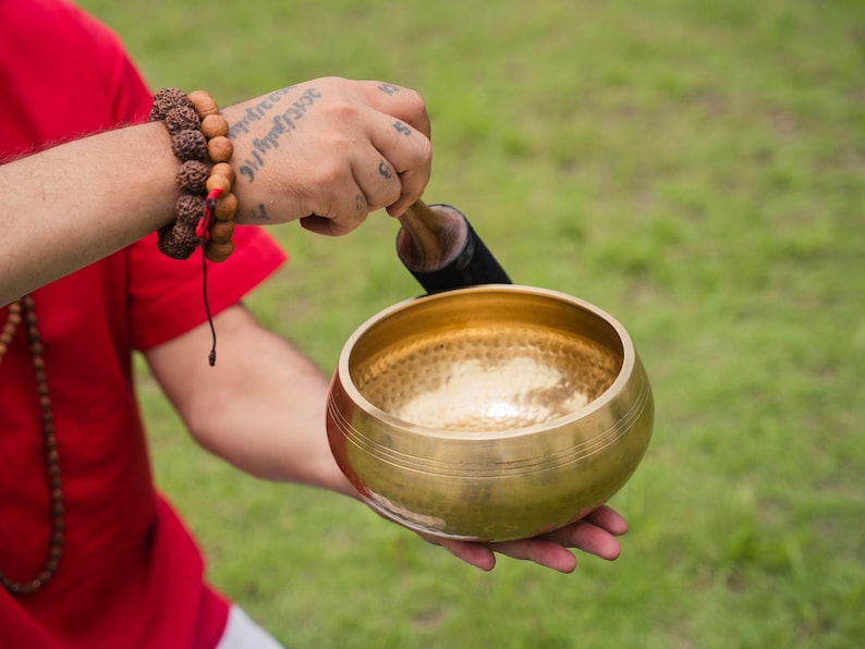 May include: A golden singing bowl with a wooden striker is held in a person's hand. The bowl has a textured interior and a smooth, polished exterior. A beaded bracelet is worn on the wrist. The background is blurred green grass.