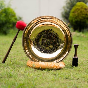 May include: A large, golden-colored gong with a mallet and a striker, resting on a decorative base. The mallet has a red head and a dark wooden handle. The striker is black with a wooden handle. The gong is set on green grass.