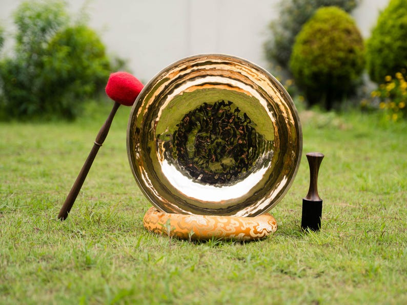 May include: A large, golden-colored gong with a mallet and a striker, resting on a decorative base. The mallet has a red head and a dark wooden handle. The striker is black with a wooden handle. The gong is set on green grass.