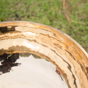 May include: Close-up of a golden singing bowl with a polished, reflective surface. The bowl's rim and interior reflect the surrounding environment, creating a rippled effect. The background is blurred green grass.