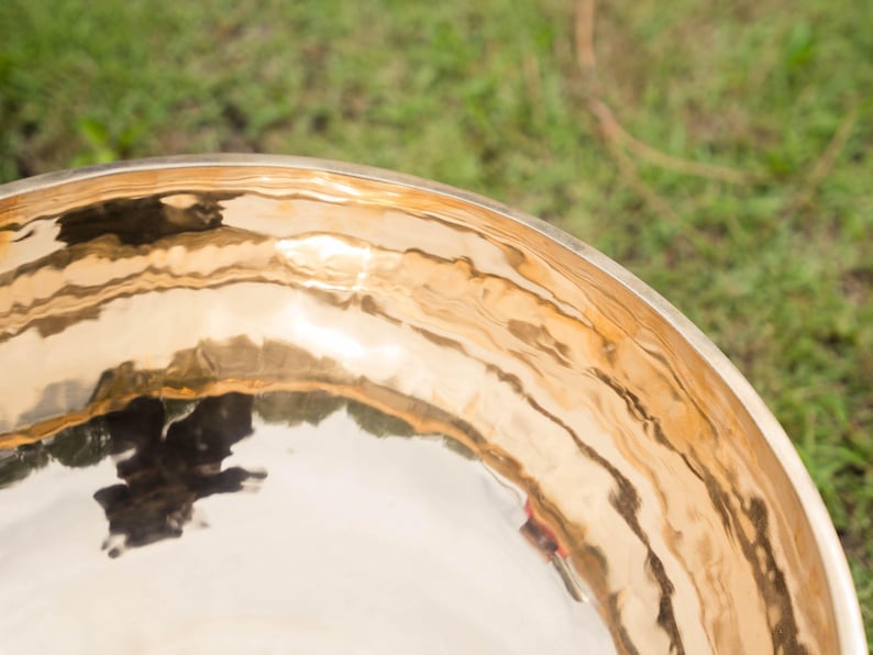 May include: Close-up of a golden singing bowl with a polished, reflective surface. The bowl's rim and interior reflect the surrounding environment, creating a rippled effect. The background is blurred green grass.