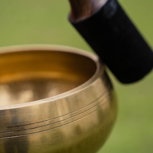 May include: Close-up of a golden singing bowl with three etched lines. A black mallet with a wooden handle rests above the bowl, ready to strike. The background is a soft green, suggesting an outdoor setting.