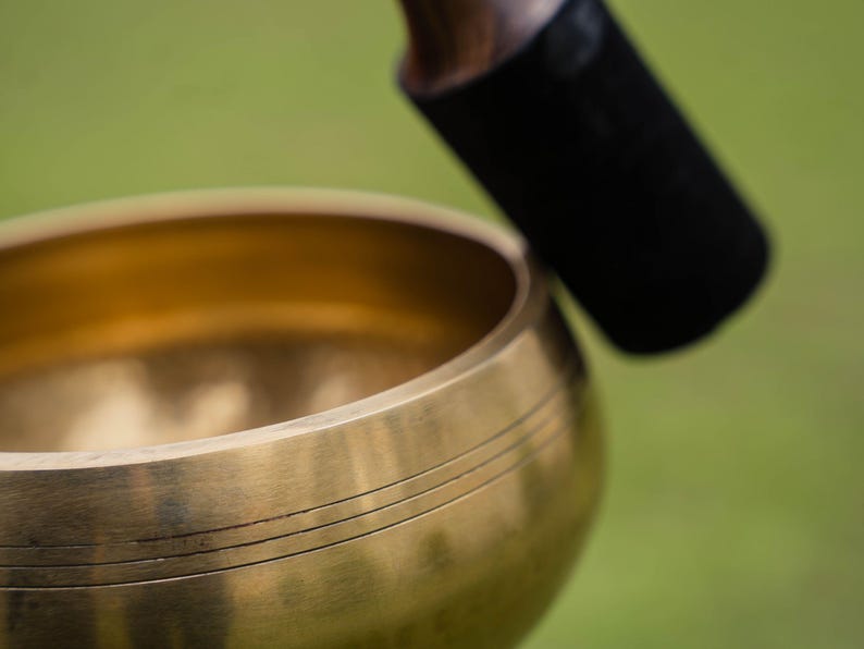 May include: Close-up of a golden singing bowl with three etched lines. A black mallet with a wooden handle rests above the bowl, ready to strike. The background is a soft green, suggesting an outdoor setting.