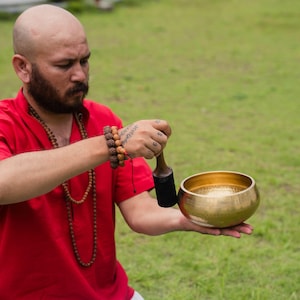 May include: A person in a red shirt holds a golden singing bowl and a wooden mallet. The bowl is round with a smooth, polished surface. The person is wearing a beaded necklace and bracelets.