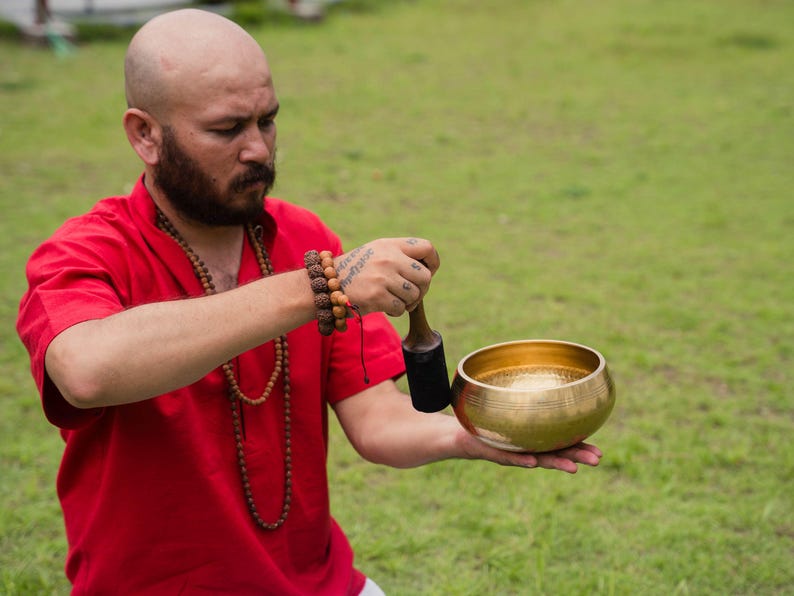 May include: A person in a red shirt holds a golden singing bowl and a wooden mallet. The bowl is round with a smooth, polished surface. The person is wearing a beaded necklace and bracelets.