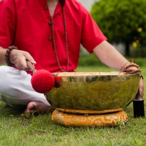 May include: A person seated on grass with a large, golden singing bowl. The bowl rests on a decorative cushion. A red mallet is poised to strike the bowl. The person wears a red shirt and white pants.