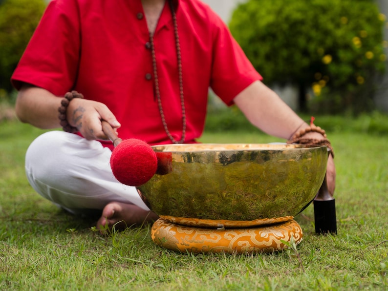 May include: A person seated on grass with a large, golden singing bowl. The bowl rests on a decorative cushion. A red mallet is poised to strike the bowl. The person wears a red shirt and white pants.