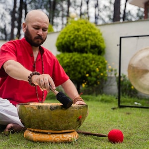 May include: A person in a red shirt and white pants is using a mallet to strike a brass singing bowl. A large gong hangs in the background. The scene takes place outdoors on a grassy lawn.