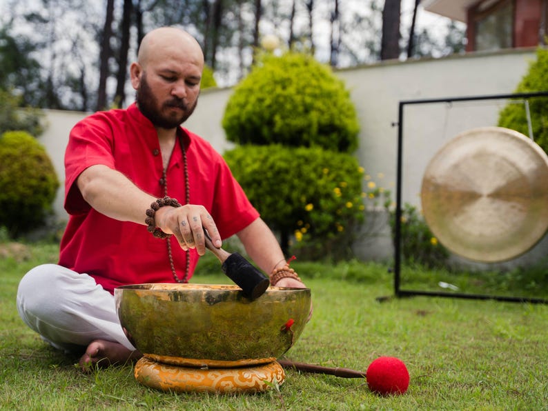 May include: A person in a red shirt and white pants is using a mallet to strike a brass singing bowl. A large gong hangs in the background. The scene takes place outdoors on a grassy lawn.