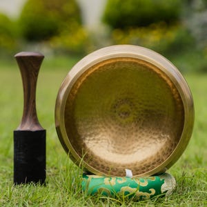 May include: A golden singing bowl with a wooden mallet and a green cushion, set on a grassy surface. The bowl has a hammered texture and is positioned next to the mallet, which has a dark wooden handle and a black base. The cushion has a gold pattern.