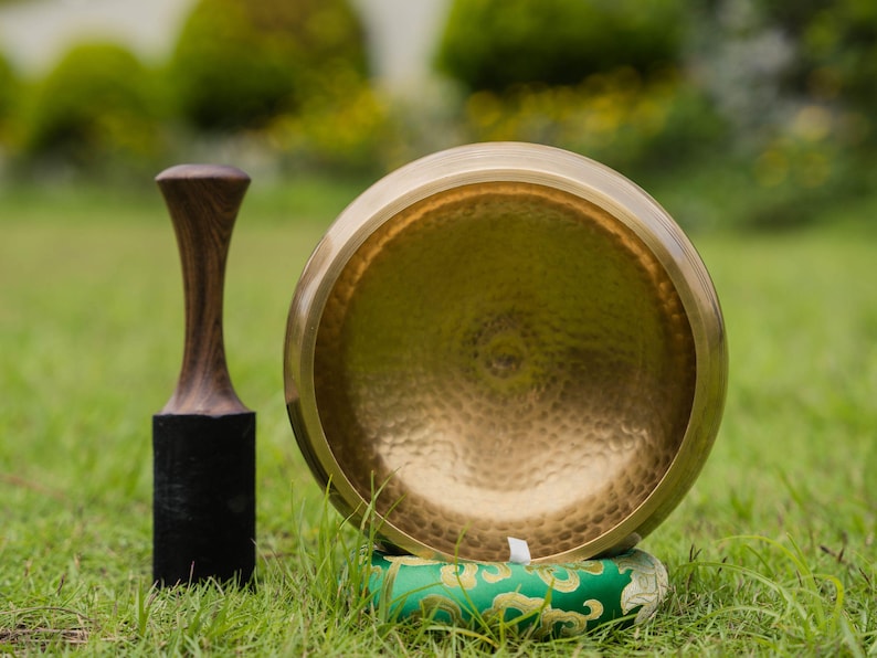 May include: A golden singing bowl with a wooden mallet and a green cushion, set on a grassy surface. The bowl has a hammered texture and is positioned next to the mallet, which has a dark wooden handle and a black base. The cushion has a gold pattern.