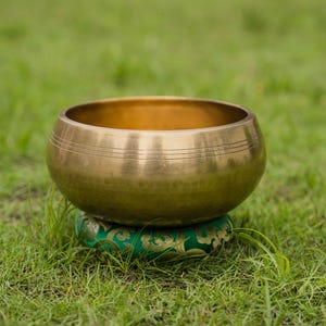 May include: A golden singing bowl with etched lines rests on a green patterned cushion, set against a backdrop of green grass. The bowl's metallic surface reflects the light, highlighting its rounded shape.